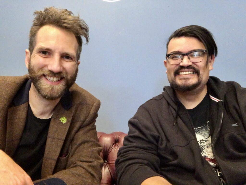 Photo of Dez (on the left) and Gavin (on the right) sat on a sofa and grinning at the camera.